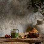 A 70g jar of Abida Rasheed traditional Malabar Garam Masala displayed on a rustic wooden table with a bowl of ground spice powder and whole authentic Kerala spices.   